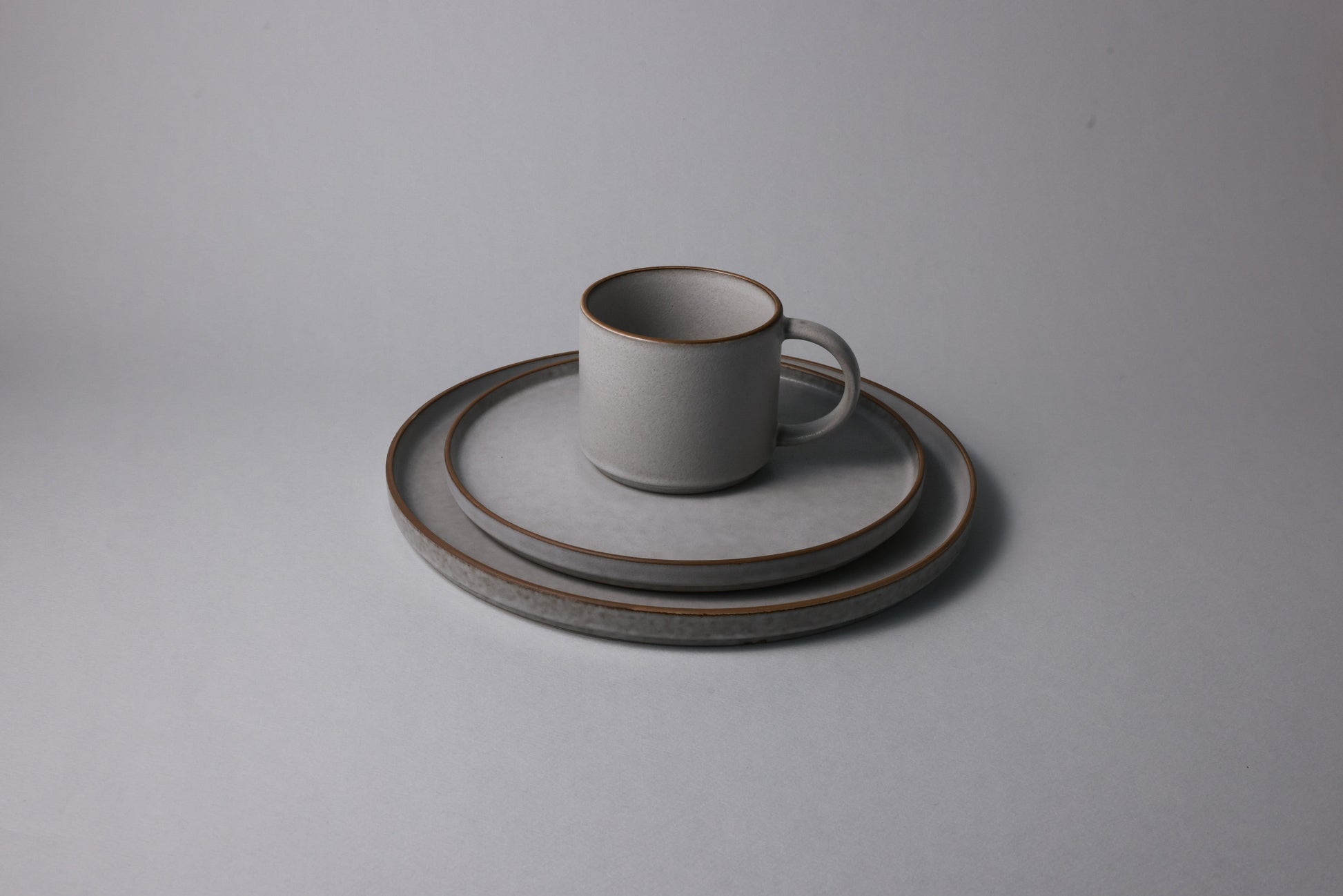 Grey stoneware mug set on stacked medium and large plates, finished with a matte glaze and subtle tonal variations, photographed against a soft neutral backdrop.