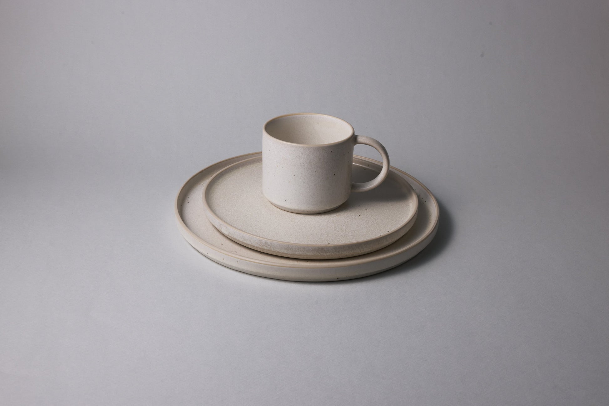 Beige stoneware mug placed on stacked medium and large plates, featuring a matte speckled finish and softly rounded edges, photographed on a neutral background.