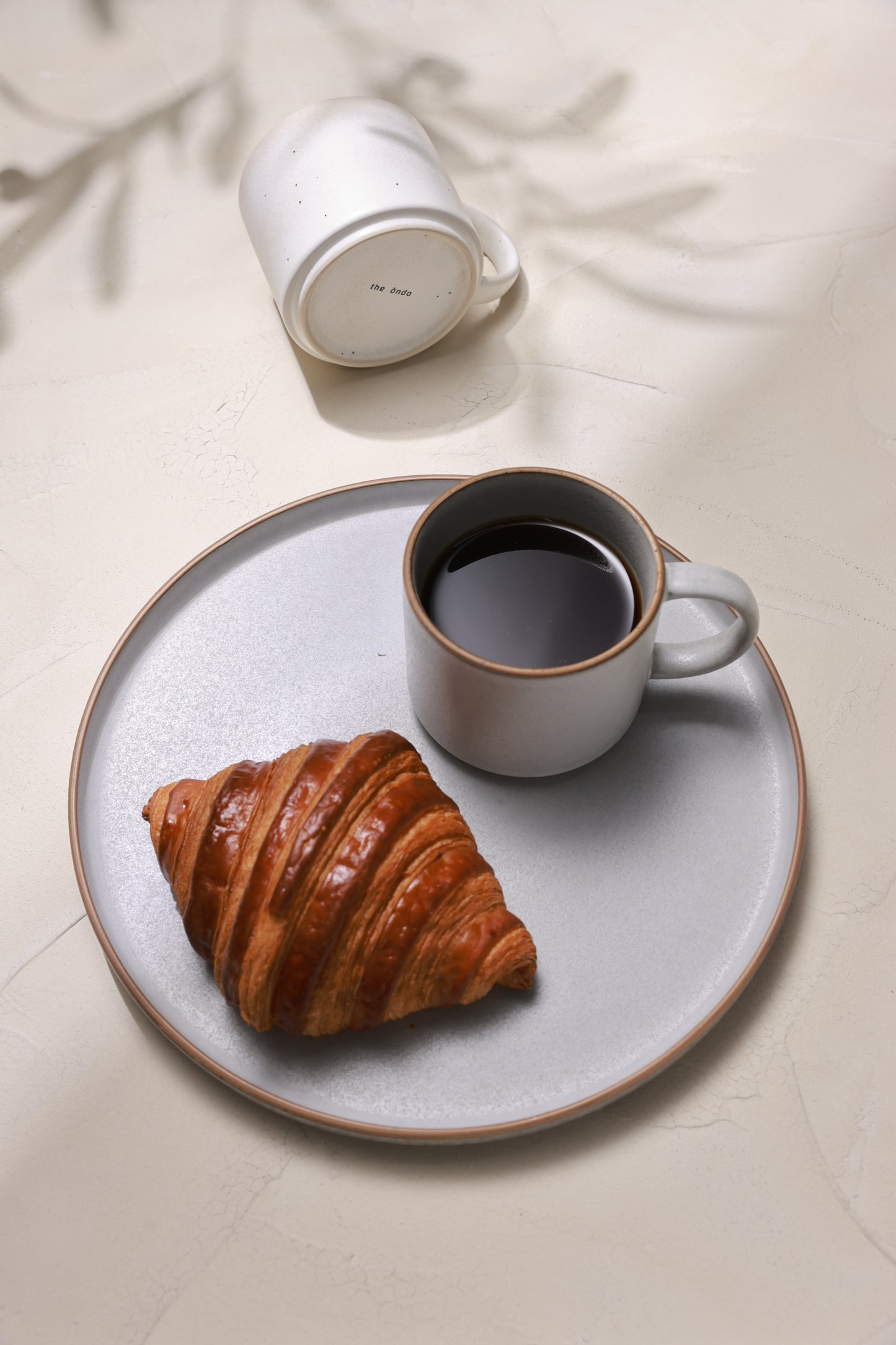 A warm coffee in a matte stoneware mug, croissant on a stoneware plate, photographed in natural light